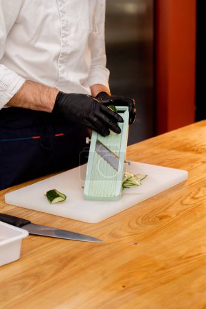 Photo for Close-up of hands in black gloves slicing cucumbers with a grater on a white board - Royalty Free Image