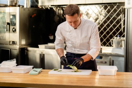 Foto de Un chef con una chaqueta blanca en la cocina con guantes negros corta pepinos antes de cocinar - Imagen libre de derechos