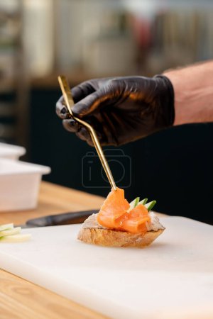 Photo for Close-up of hands in black gloves placing a piece of salmon on bread with golden tweezers - Royalty Free Image