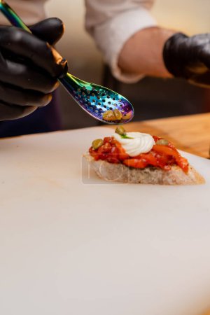 Foto de Primer plano de chefs manos en guantes negros cucharadas alcaparras en sándwich de pimienta - Imagen libre de derechos