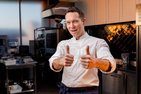 Foto de Cocina profesional sonriente y satisfecho chef en una bata blanca se para con los dedos levantados - Imagen libre de derechos