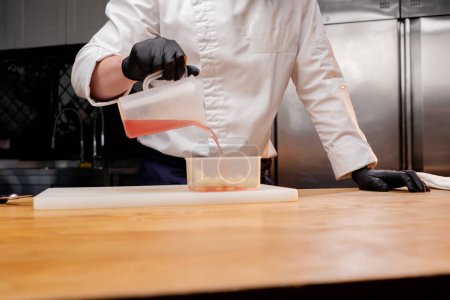 Téléchargez les photos : Un chef dans une veste blanche dans une cuisine professionnelle verse la sauce finie dans un récipient avec des morceaux de pommes rondes - en image libre de droit