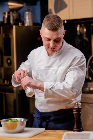 Nahaufnahme eines Kochs in weißer Uniform in einer professionellen Küche, der bereit ist, einen Salat zu salzen