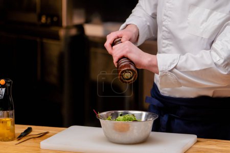 Nahaufnahme eines Kochs in weißer Uniform in einer professionellen Küche, der einen Salat in einer Metallschüssel salzt