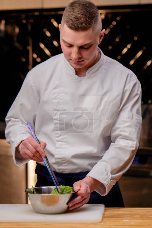 Ein Koch in weißer Uniform in einer professionellen Küche rührt mit einer großen Pinzette einen Salat um