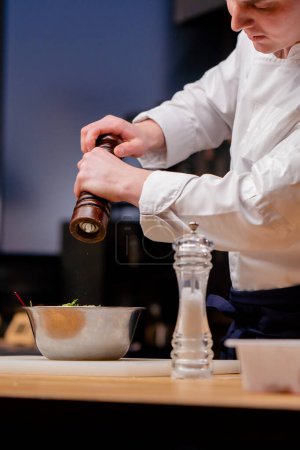 Ein Koch in weißer Uniform in einer professionellen Küche salzt einen Salat in einer Metallschüssel