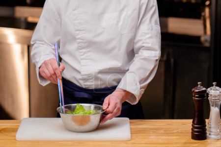 Photo for Close-up of a chef in a white uniform in a professional kitchen stirring a salad with large tweezers - Royalty Free Image