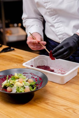 Foto de Primer plano de un plato negro en el que la ensalada las manos de los chefs le agregan trozos de remolacha con pinzas - Imagen libre de derechos