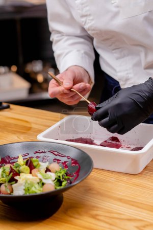 close-up of a black plate in which salad the chefs hands add pieces of beet to it with tweezers