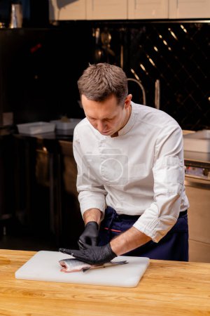 Foto de Un chef con una chaqueta blanca en una cocina profesional corta pescado con un cuchillo antes de cocinar - Imagen libre de derechos
