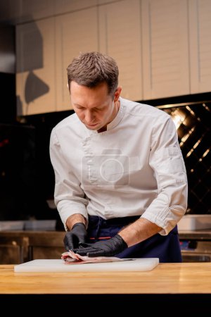 Foto de Un chef con una chaqueta blanca en una cocina profesional corta pescado con un cuchillo antes de cocinar - Imagen libre de derechos