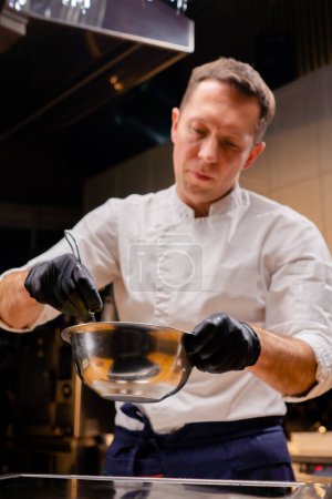 Foto de Un chef con una chaqueta blanca en la cocina agita el contenido en una sartén con un batidor mientras lo sostiene en sus manos - Imagen libre de derechos