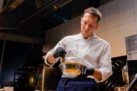 Téléchargez les photos : Un chef vêtu d'une veste blanche dans la cuisine remue le contenu dans une casserole avec un fouet tout en le tenant dans ses mains - en image libre de droit