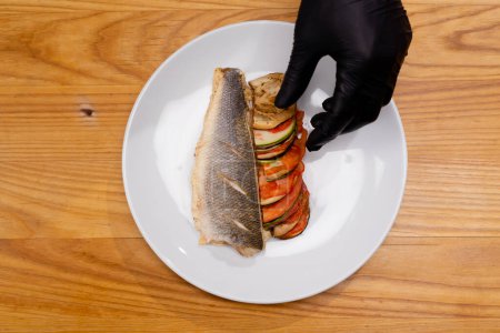 Photo for Top close-up frame of hands and cooked fish dish and grilled vegetables on a white plate - Royalty Free Image