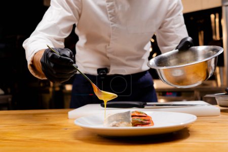 Photo for Close-up of a chefs hands holding a bowl and pouring sauce from a spoon onto the finished dish - Royalty Free Image