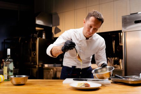 Téléchargez les photos : Chef dans une tunique blanche tenant un bol et verser la sauce d'une cuillère sur le plat fini - en image libre de droit