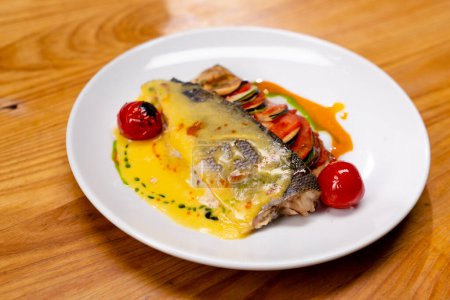 Téléchargez les photos : Gros plan d'une assiette blanche sur la table avec un plat de poisson préparé avec des légumes grillés et deux tomates - en image libre de droit