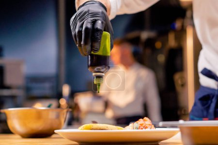 close-up of a chefs hands pouring green sauce from a bottle onto a finished dish