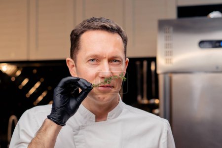 Téléchargez les photos : Chef visage dans une cuisine professionnelle dans une veste blanche se tient et renifle un brin de romarin - en image libre de droit