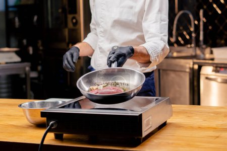 Téléchargez les photos : Gros plan du chef tenant une poêle avec un morceau de poitrine de canard rose avec du romarin sur la cuisinière - en image libre de droit