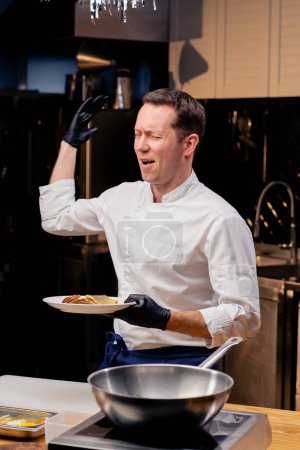 Foto de Un chef en una cocina profesional se pone de pie con un plato blanco de un plato de pescado listo se regocija y baila - Imagen libre de derechos