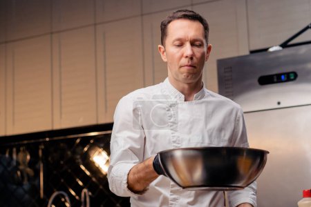 Foto de Chef en una cocina profesional se para con una sartén en la mano y lanza el contenido al aire - Imagen libre de derechos