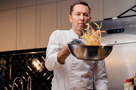 Téléchargez les photos : Chef dans une cuisine professionnelle se tient avec une poêle à frire à la main et jette le contenu dans l'air - en image libre de droit