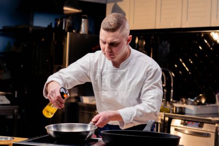 Téléchargez les photos : Un chef dans un uniforme blanc pulvérise une poêle à frire avec de l'huile d'un vaporisateur dans une cuisine professionnelle - en image libre de droit