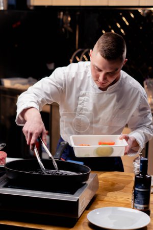 Téléchargez les photos : Un chef en uniforme blanc dans une cuisine professionnelle met des morceaux de légumes sur une poêle à frire avec pinces - en image libre de droit
