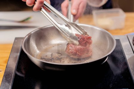 Foto de Primer plano en una cocina profesional con pinzas que sostienen un filete de ternera sobre una sartén caliente con aceite - Imagen libre de derechos