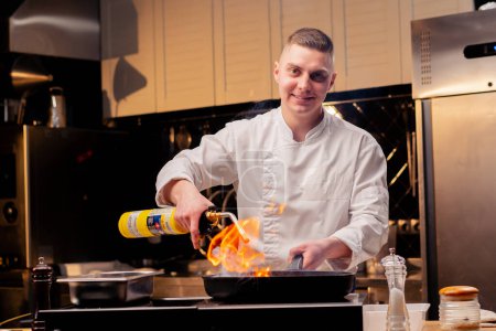Foto de Cocinero sonriente freír diferentes verduras en una sartén caliente con una antorcha en la estufa - Imagen libre de derechos