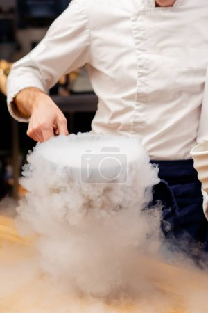 Foto de Primer plano de un chef en un uniforme blanco en la cocina preparando nitrógeno líquido que se extiende por toda la mesa - Imagen libre de derechos