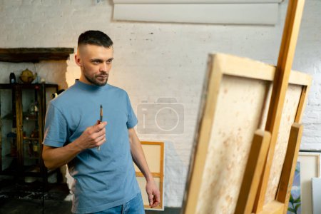 Téléchargez les photos : Dans un atelier d'art un artiste en t-shirt bleu réfléchit à l'endroit où faire un trait avec un couteau de palette - en image libre de droit