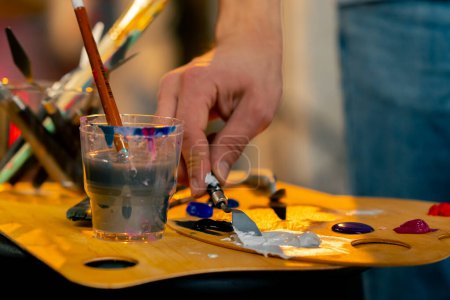 Foto de Primer plano en un taller de arte un artista en camiseta azul utiliza un cuchillo de paleta para amasar el color blanco en una paleta - Imagen libre de derechos