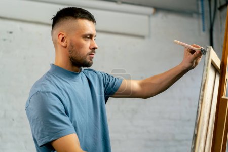 Foto de En un taller de arte un artista en una camiseta azul hace grandes movimientos en el lienzo con un cepillo ancho y plano - Imagen libre de derechos