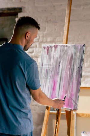 Foto de En un taller de arte un artista en camiseta azul dibuja sobre un lienzo con un cuchillo de paleta - Imagen libre de derechos