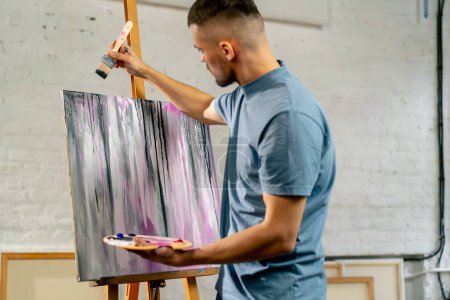 Téléchargez les photos : Dans un atelier d'art, l'artiste crée des stries sombres sur la toile avec un grand pinceau - en image libre de droit