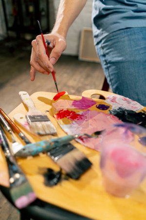 Photo for Close up in an art studio an artist sitting dips a brush in red paint on a palette - Royalty Free Image