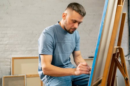 Photo for Young artist in a blue t-shirt in an art studio working on a painting while sitting - Royalty Free Image
