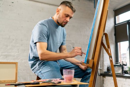 Téléchargez les photos : Jeune artiste en t-shirt bleu dans un atelier d'art travaillant sur une peinture assis - en image libre de droit