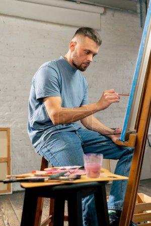 Foto de Joven artista en una camiseta azul en un estudio de arte trabajando en una pintura mientras está sentado - Imagen libre de derechos