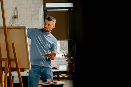 Téléchargez les photos : Général tourné jeune artiste dans un t-shirt bleu dans un atelier d'art travaillant sur une peinture - en image libre de droit