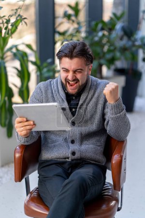 Téléchargez les photos : Un jeune employé de bureau sur fond de fenêtre assis sur une chaise travaillant avec une tablette sentiment de vainqueur - en image libre de droit