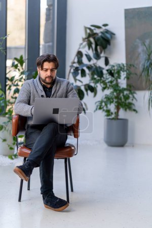 Téléchargez les photos : D'un jeune employé de bureau sur fond de fenêtre assis sur une chaise travaillant sur un ordinateur portable - en image libre de droit