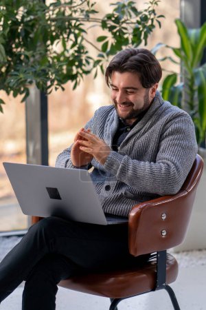 Téléchargez les photos : Jeune gars employé de bureau sur le fond d'une fenêtre assis sur une chaise travaillant un appel de travail ordinateur portable - en image libre de droit