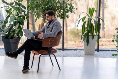 Téléchargez les photos : Jeune gars employé de bureau sur le fond d'une fenêtre assis sur une chaise travaillant un appel de travail ordinateur portable - en image libre de droit
