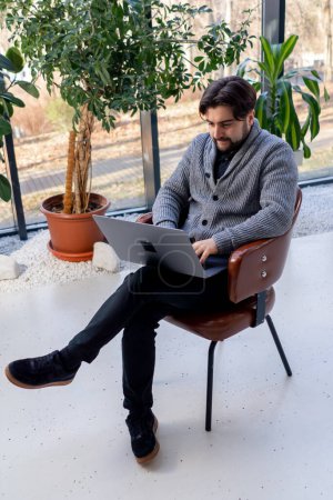 Foto de De un joven trabajador de oficina sobre el fondo de una ventana sentada en una silla trabajando en un ordenador portátil - Imagen libre de derechos