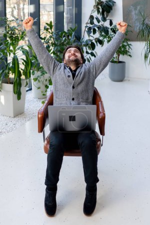Foto de Un joven trabajador de oficina en el fondo de una ventana sentado en una silla de trabajo con una sensación de tableta de vencedor - Imagen libre de derechos