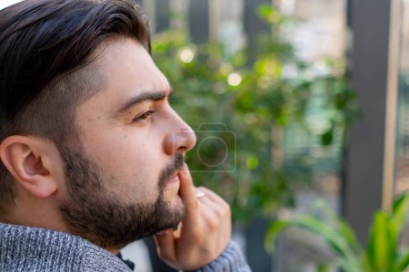 Nahaufnahme eines jungen Mannes mit Bart, der nachdenklich aus dem Fenster zwischen den Pflanzen blickt