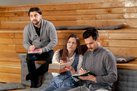 Téléchargez les photos : Trois employés de bureau assis dans le salon sur des bancs en bois avec des oreillers étudiant l'écriture dans un cahier - en image libre de droit